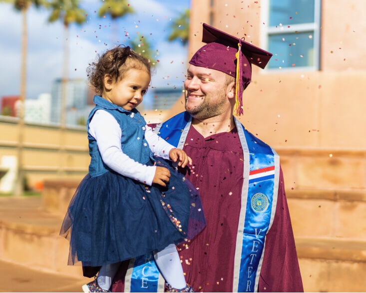 An ASU在线 graduate dressed in a cap gown holds his daughter and smiles while confetti falls around them.