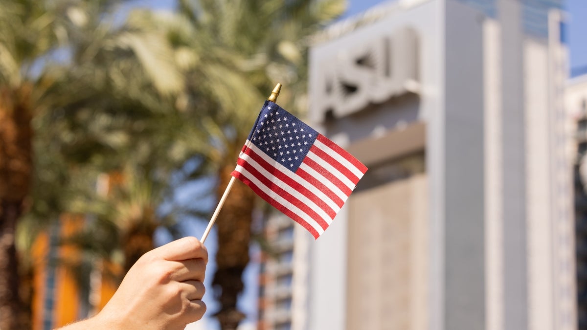 A hand holds a tiny American flag in front of an ASU sign